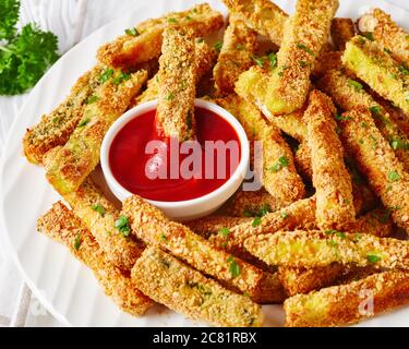 close-up of crunchy zucchini sticks breaded with panko breadcrumbs, parmesan cheese, spices on a white plate with ketchup on a wooden table, macro Stock Photo