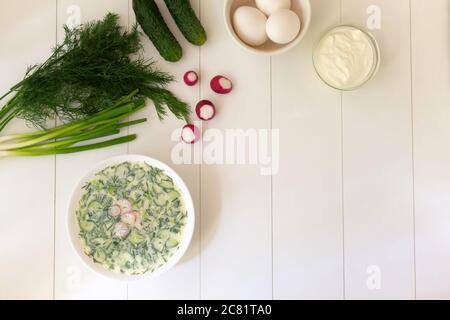 Cold soup in a deep white plate with ingredients on a white table. Okroshka, a traditional dish of Russian cuisine. Copy space. Top view. Stock Photo
