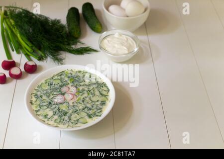 Cold soup in a deep white plate with ingredients on a white table. Okroshka, a traditional dish of Russian cuisine. Copy space. Stock Photo