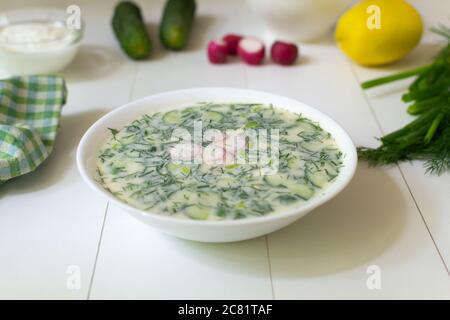 Cold soup in a deep white plate with ingredients on a white table. Okroshka, a traditional dish of Russian cuisine. Selective focus. Stock Photo