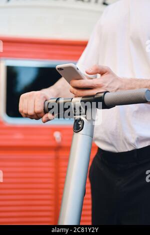 Close up young man standing with electric scooter and cellphone in hand on street Stock Photo