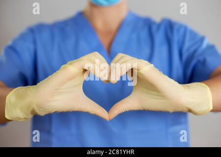 Doctor or nurse in a blue shirt makes the heart symbol with hands in gloves. Stock Photo