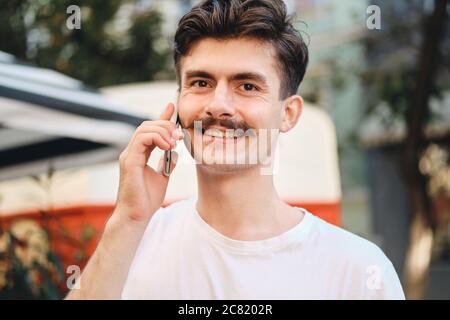 Portrait of young smiling mustache man in withe T-shirt talking on cellphone while happily looking in camera on city street Stock Photo