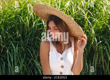 Happy stylish woman sitting in front of grass and looking while holding her hat Stock Photo