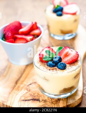 homemade, exquisite dessert tiramisu in glasses decorated with strawberry, blueberry, mint on white wooden table, top view. Stock Photo