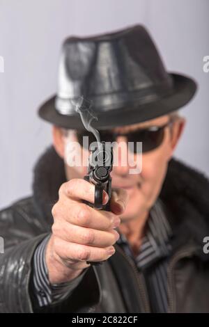 Elderly man with jacket, hat, and black glasses aiming straight ahead with gun from which smoke comes out (Select focus) Stock Photo