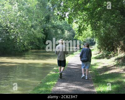 A man and woman in summer clothing walk along the towpath in dappled sunlight under the trees at the side of the Grand Union Canal Stock Photo