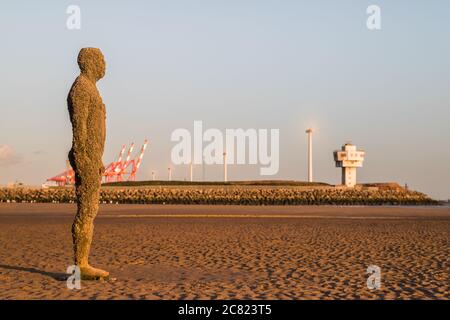 An Iron Man statue on seen on the beach at Crosby near Liverpool (England) in July 2020 in front of a row of spinning wind turbines in Liverpool dock. Stock Photo