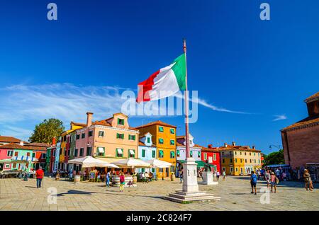 Burano, Italy, September 14, 2019: Burano island central town square with old colorful buildings and waving italian flag, blue sky in sunny summer day background, Venice Province, Veneto Region