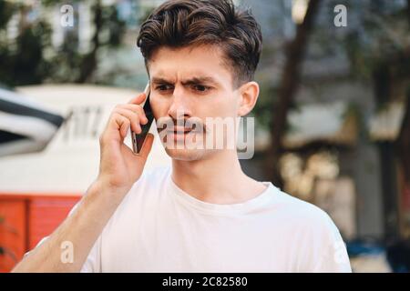 Portrait of young amazed mustache man in withe T-shirt talking on cellphone on street Stock Photo