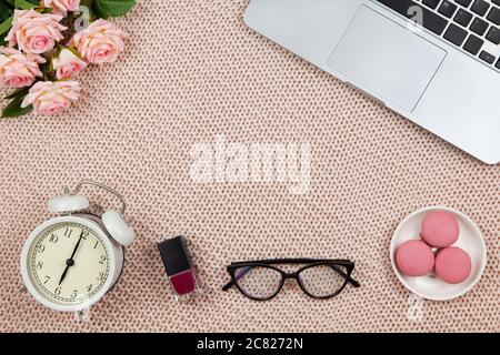 Modern female working space, top view. Laptop, glasses, nail polish, cakes, roses, clock on knitted blanket, copy space, flat lay. Desktop of freelanc Stock Photo