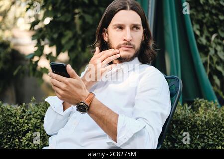 Young confident brunette man in shirt with cellphone thoughtfully looking away outdoor Stock Photo