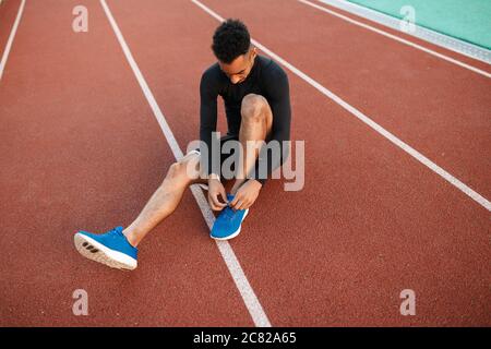 Young African American sporty man tying shoelaces on sneakers on racetrack at city stadium Stock Photo