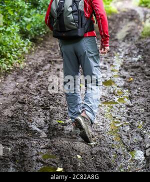 Rear view, low section of isolated man walking with backpack on muddy woodland path in UK countryside. Rambler from behind in mud in rural setting. Stock Photo