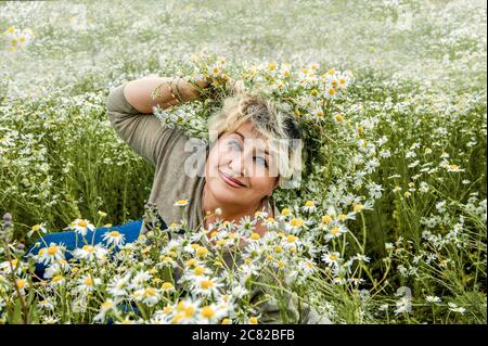 Mature beautiful woman with a wreath and a bouquet of wild flowers in a large field of daisies. Active recreation, perfect maturity. Stock Photo