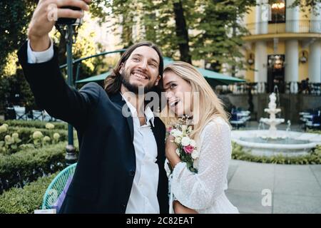 Beautiful romantic newlyweds happily taking selfie on cellphone in garden of restaurant Stock Photo