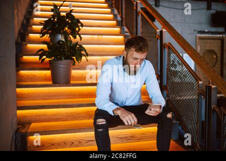 A young caucasian man sitting on the stairs in a cafe and typing a message on the phone. Handsome businessman in a restaurant Stock Photo