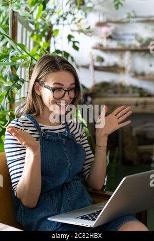 Woman gardener in glasses wear blue jeans overalls, sitting on chair in greenhouse, resting, using laptop after work, smiling and speaking on video ca Stock Photo