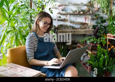 Woman gardener in glasses wear blue jeans overalls, sitting on chair in greenhouse, using laptop after work, communicates on internet with customer su Stock Photo