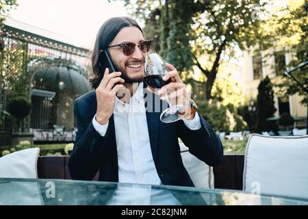 Young handsome smiling brunette bearded man happily talking on cellphone and drinking wine while resting in restaurant outdoor Stock Photo