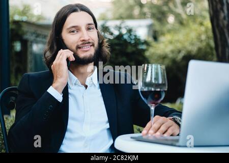 Young confident bearded businessman with glass of wine happily looking in camera while talking on cellphone in restaurant outdoor Stock Photo