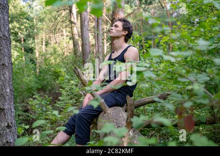 Side view of thoughtful man looking away while sitting on log against trees in forest Stock Photo