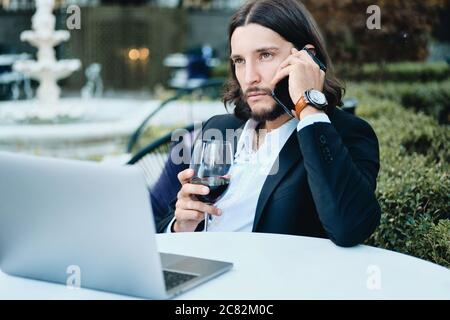 Young serious confident bearded businessman with glass of wine talking on cellphone in restaurant outdoor Stock Photo