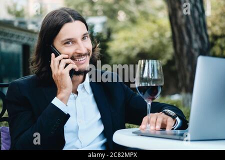 Young handsome brunette bearded businessman with glass of wine happily looking in camera while talking on cellphone in restaurant outdoor Stock Photo