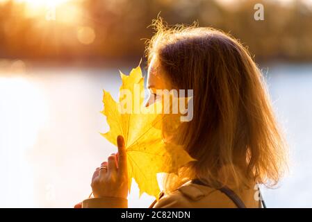 Autumn maple leaf in hand of pretty woman, portrait outside in autumn season. Beautiful fall nature, scene by river in autumn park against sun, young Stock Photo