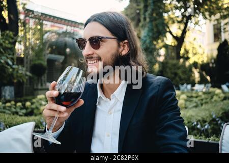 Young handsome smiling brunette bearded man happily drinking wine in restaurant outdoor Stock Photo