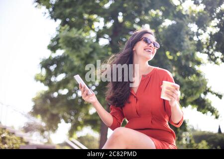 Young smiling pretty woman drinking coffee while reading messages on the mobile phone Stock Photo