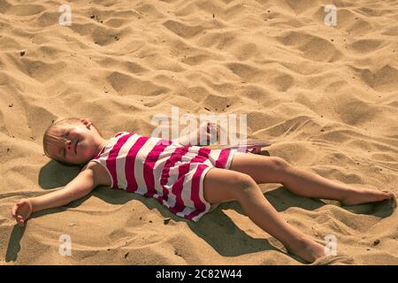 Baby girl lying on sand. Adorable kid sunbathing at summertime. Young child resting and relaxing with her eyes closed. Caucasian preschooler lying dow Stock Photo