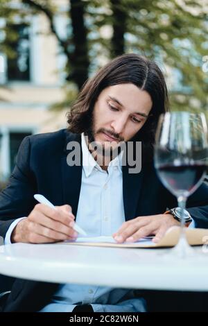 Young attractive bearded businessman with glass of red wine thoughtfully working in restaurant outdoor Stock Photo