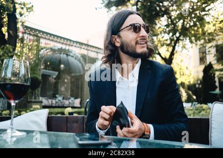 Young handsome bearded man happily looking away with wallet in hand in restaurant outdoor