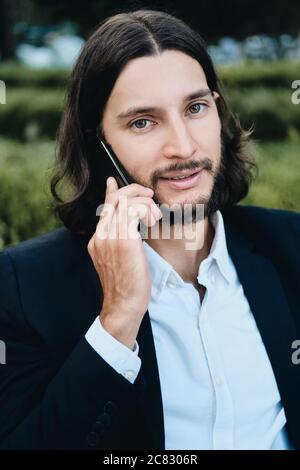 Young attractive confident bearded businessman looking in camera while talking on cellphone in restaurant outdoor Stock Photo