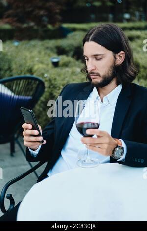 Young confident bearded businessman with glass of red wine thoughtfully using cellphone in restaurant outdoor Stock Photo