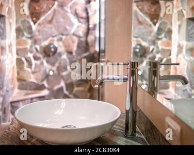 Close up view of a single white ceramic basin, polished chrome faucet, and mirror in a luxury hotel bathroom with a natural stone shower stall. Stock Photo