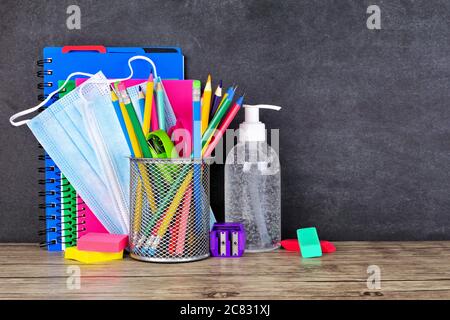School supplies and COVID 19 prevention items on a desk against a chalkboard background. Back to school during pandemic concept. Stock Photo