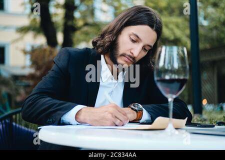 Young handsome serious bearded businessman with glass of red wine thoughtfully working in restaurant outdoor Stock Photo