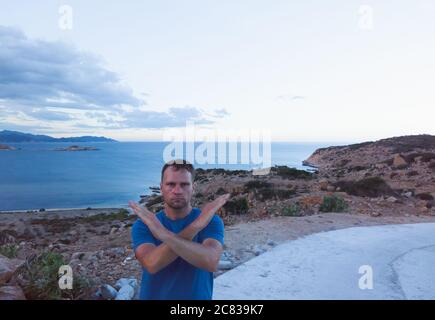 Serious man standing with crossed hands on a sunset mountain rock and sea background. Stop vacation, travel because of coronavirus or be careful in Stock Photo