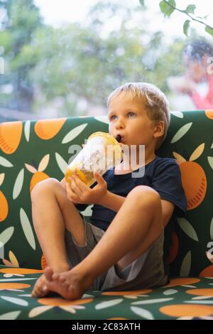 Front view of happy thirsty blond kid boy drinking lemonade, fruit tea, kombucha ice tea with peach and lemon, wellness orange drinks from plastic cup Stock Photo