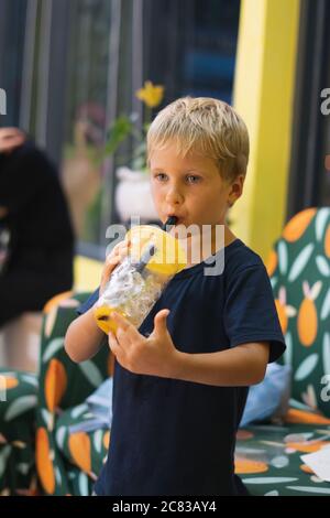 Front view of happy thirsty blond kid boy drinking lemonade, fruit tea, kombucha ice tea with peach and lemon, wellness orange drinks from plastic cup Stock Photo