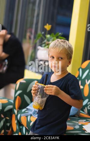 Front view of happy thirsty blond kid boy drinking lemonade, fruit tea, kombucha ice tea with peach and lemon, wellness orange drinks from plastic cup Stock Photo