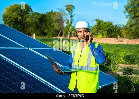 young male technician worker wearing green vests and helmet with laptop in hand talking on phone about the maintenance and installation of the solar p Stock Photo