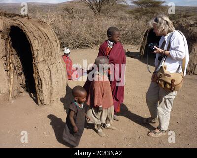 Female tourist talks to Masai children near village mud huts, Tanzania Stock Photo