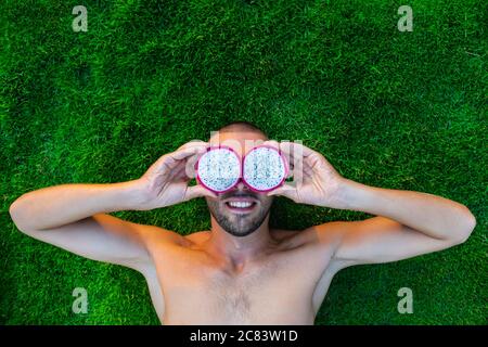 Smiling young man lying on a green grass with dragon fruit instead of his eyes Stock Photo