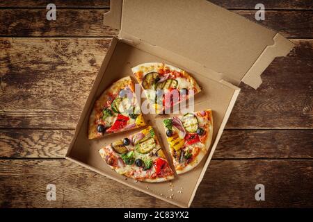 Sliced Italian pizza with fresh vegetables and salami in a cardboard fast food box for delivery on a rustic wooden table in a top down view Stock Photo