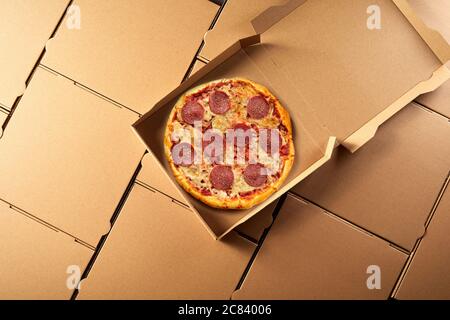 Open takeaway box with freshly made Italian salami pizza inside on a background of generic brown cardboard pizza boxes viewed top down in a flat lay s Stock Photo