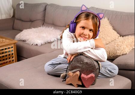 Little girl in musical headphones and a backpack is ready to go to school. A European schoolgirl demonstrates positive emotions. Education and school days concept. Copy space Stock Photo