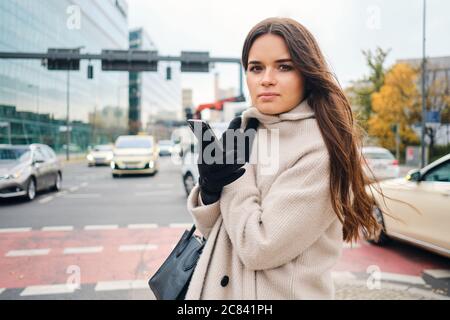 Attractive stylish brunette girl in coat with cellphone intently looking in camera walking around street Stock Photo